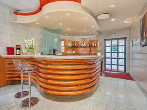 Curved wooden bar with marble countertop, transparent bar stools, bottles of alcohol, plants, and entrance door at B&B Hotel Hotel Prestige Ornago