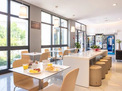 Dining area at B&B Hotel Mantova with tables and chairs, breakfast food on the table, large windows, and vending machines in the background