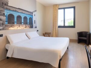 Double bed with white linens below a large mural next to a window with yellow patterned curtains at B&B Hotel Mantova
