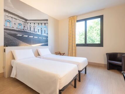 Two single beds with white linens and pillows under a large black and white historic building photo, window with yellow curtains in B&B Hotel Mantova