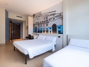 Two beds with white linens, a wooden chair, desk, and large black and white architectural print in a room at B&B Hotel Mantova