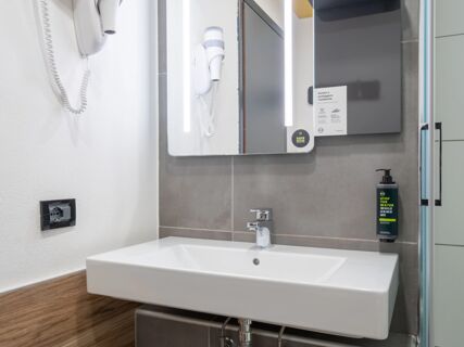 White rectangular sink with a faucet under a mirror with LED lights, wall-mounted hairdryer, and shampoo dispenser at B&B Hotel Mantova