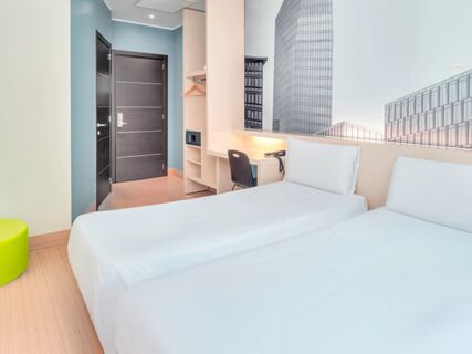 Two single beds with white linens face a desk and chair with a phone, open closet with hangers and electronic safe in a room at B&B Hotel Milano Cenisio Garibaldi