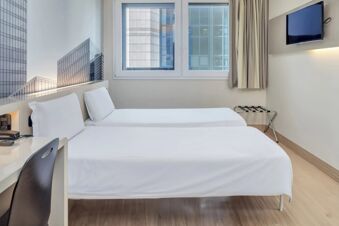 Hotel room at B&B Hotel Milano Cenisio Garibaldi with two single beds, a window showing tall buildings, a mounted TV and a small desk with chair