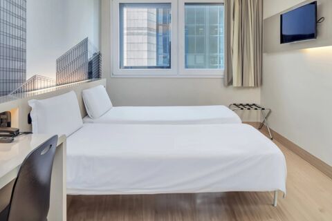 Hotel room at B&B Hotel Milano Cenisio Garibaldi with two single beds, a window showing tall buildings, a mounted TV and a small desk with chair