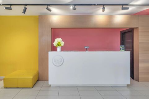 Reception area with a white front desk, white flowers in a vase, yellow seating, pink wall, and B&B Hotel Milano Cenisio Garibaldi logo