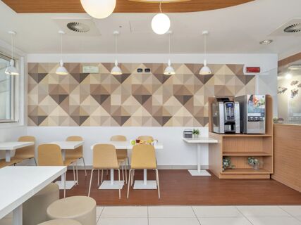 Dining area with wooden chairs, white tables, geometric patterned wall, and coffee machines at B&B Hotel Milano Cenisio Garibaldi