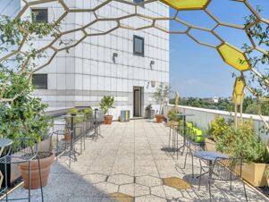 Outdoor terrace at B&B Hotel Milano Cenisio Garibaldi featuring metal chairs, tables, potted plants, and a yellow geometric frame with sky background