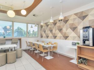 Dining area with white tables, light wood chairs, cylindrical stools, pendant lights, geometric wall pattern, and coffee machine at B&B Hotel Milano Cenisio Garibaldi