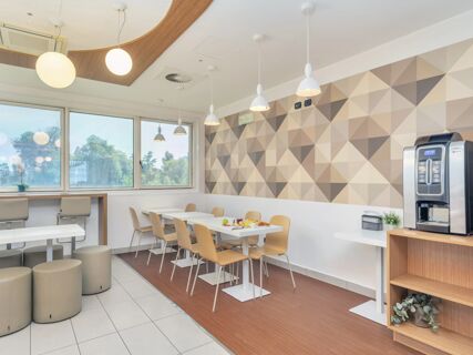 Dining area with white tables, light wood chairs, cylindrical stools, pendant lights, geometric wall pattern, and coffee machine at B&B Hotel Milano Cenisio Garibaldi