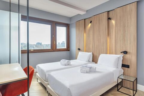 Two single beds with white linens and towels, wooden headboard with spotlights, window, desk, and red chair in B&B Hotel Milano City Center Duomo room