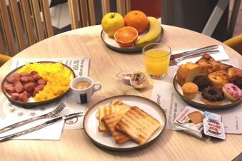 A breakfast spread at B&B Hotel Milano City Center Duomo with scrambled eggs, sausage, toast, croissants, muffins, fruit plate, orange juice, and espresso coffee on a round wooden table