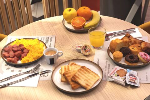 A breakfast spread at B&B Hotel Milano City Center Duomo with scrambled eggs, sausage, toast, croissants, muffins, fruit plate, orange juice, and espresso coffee on a round wooden table