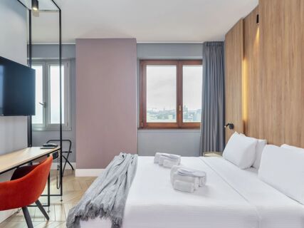 Modern hotel room at B&B Hotel Milano City Center Duomo with a double bed, gray throw, orange chair, and mounted TV on a wooden desk