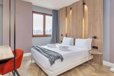 Double bed with white bedding and gray throw blanket against a light wooden headboard with reading lamps at B&B Hotel Milano City Center Duomo
