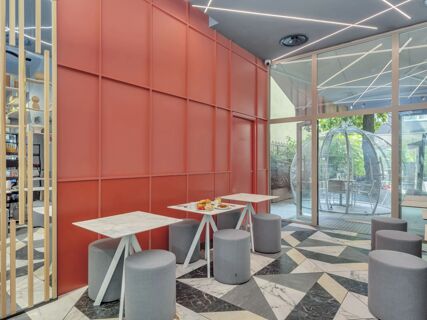 Interior seating area with red paneled wall, gray padded stools, marble tables, and large glass doors at B&B Hotel Milano City Center Duomo