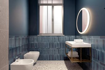 Bathroom interior at B&B Hotel Milano Duomo Velasca with blue tiled walls, white bidet and toilet, window, and illuminated round mirror