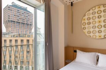 Open window with balcony railing overlooking buildings including Velasca Tower seen from a B&B Hotel Milano Duomo Velasca room with bed and wall art