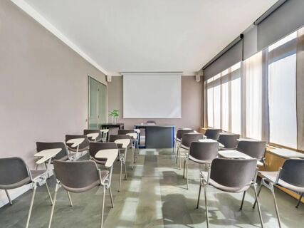 Training room with rows of grey chairs with white writing tablets, a table, and blank screen at B&B Hotel Milano La Spezia