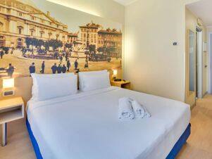 Bed with white linens and pillows in a B&B Hotel Milano La Spezia room featuring a wall mural of a historic city scene