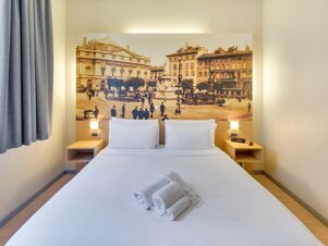 White bed with pillows and folded towels in front of a large vintage cityscape photo and bedside tables in B&B Hotel Milano La Spezia