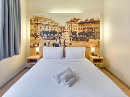 White bed with pillows and folded towels in front of a large vintage cityscape photo and bedside tables in B&B Hotel Milano La Spezia