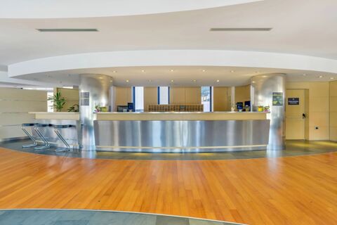 Spacious hotel lobby with a curved reception desk, metal columns, stools, plants, and wooden floors at B&B Hotel Milano La Spezia
