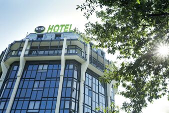 High-rise building with large glass windows and balconies under a B&B Hotel Milano San Siro sign and tree branches with sunlight shining through