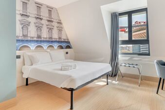 White bed with pillows and folded towels, architectural wall decor, window with gray curtains and a chair in B&B Hotel Milano San Siro
