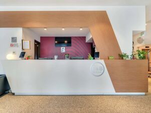Reception desk with wood paneling and white counters, two computer monitors, plants, and a TV on a red wall at B&B Hotel Milano San Siro
