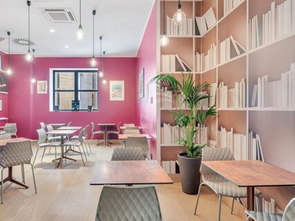Dining area at B&B Hotel Milano San Siro with pink walls, white chairs, wooden tables, hanging light bulbs, and a plant