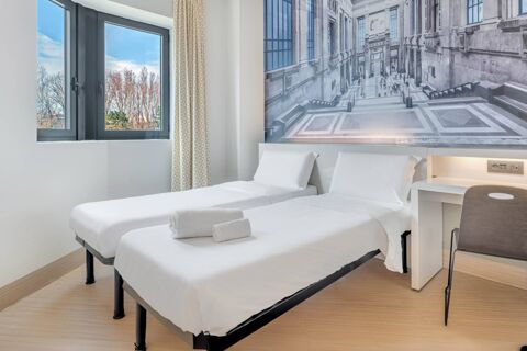 Two single beds with white linens side by side, a window with patterned curtains, and a desk in B&B Hotel Milano San Siro