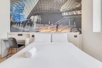 White bed with pillows and a rolled towel, desk with black chair, and a large photo mural in B&B Hotel Milano San Siro