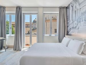 Bright hotel room at B&B Hotel Milano Sant'Ambrogio with large windows, gray curtains, white bed linens, and a cityscape mural on the wall