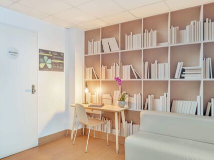 Study area with a wooden desk, chair, lamp, purple orchid, and wall-mounted shelves with white books at B&B Hotel Milano Sant'Ambrogio