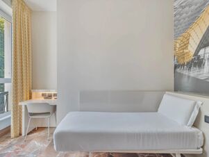 Minimalist room at B&B Hotel Milano Sant'Ambrogio with white bed, yellow patterned curtains, white chair, desk, and large window