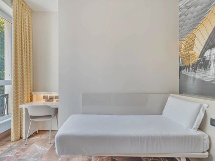 Minimalist room at B&B Hotel Milano Sant'Ambrogio with white bed, yellow patterned curtains, white chair, desk, and large window