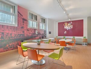 Dining area with round and oval tables surrounded by green and orange chairs under windows at B&B Hotel Milano Sant'Ambrogio