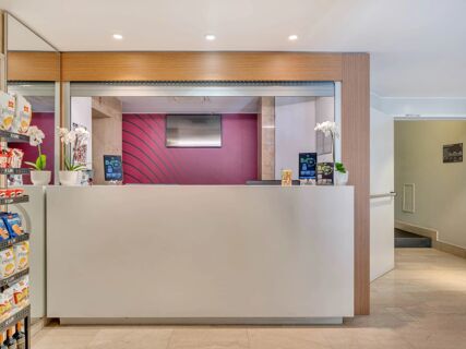 Reception desk with a white counter, digital sign displays, potted orchids, and shelves with snacks at B&B Hotel Milano Sant'Ambrogio