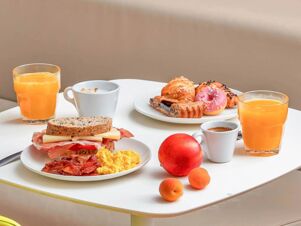 Breakfast spread at B&B Hotel Milano Sant'Ambrogio featuring scrambled eggs, bacon, sandwich, croissants, donut, coffee cups, juice, and fruits