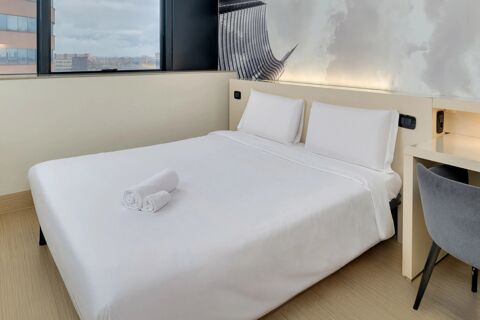 White bed with two pillows and rolled towels, city view window, desk, and wall art at B&B Hotel Milano Sesto Marelli