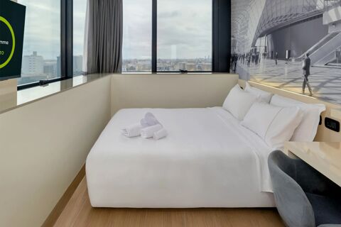 Room with large white bed, folded towels, large window with city view, wall art, TV, and chair at B&B Hotel Milano Sesto Marelli