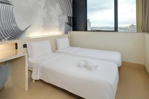 Two white single beds with pillows and rolled towels in a bright room next to a large window at B&B Hotel Milano Sesto Marelli