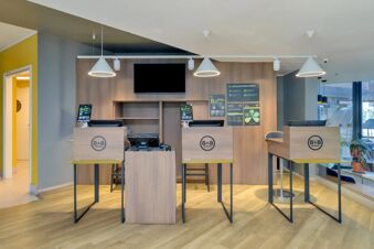 Reception area of B&B Hotel Milano Sesto Marelli with three wooden check-in desks, chairs, pendant lights, and informational posters on the wall