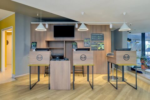 Reception area of B&B Hotel Milano Sesto Marelli with three wooden check-in desks, chairs, pendant lights, and informational posters on the wall