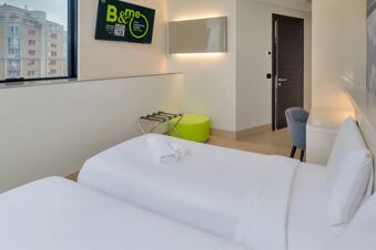 Two neatly made single beds with white linens, a wall-mounted TV, a window showing buildings, and a chair in a room of B&B Hotel Milano Sesto Marelli