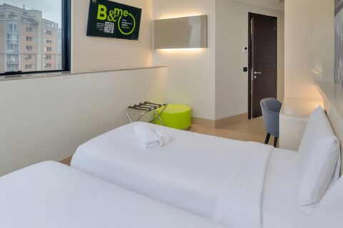 Two neatly made single beds with white linens, a wall-mounted TV, a window showing buildings, and a chair in a room of B&B Hotel Milano Sesto Marelli