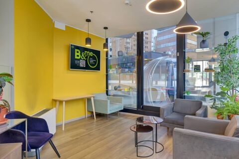 Interior view of a seating area with armchairs, small tables, hanging lights, plants, and a yellow wall with a screen at B&B Hotel Milano Sesto Marelli