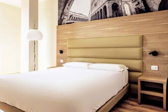 Large bed with white linens and two pillows, beige padded headboard, wooden wall panel, nightstand, and lamp at B&B Hotel Fabriano Janus