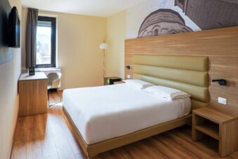 Hotel bedroom at B&B Hotel Fabriano Janus with a double bed, wooden desk, chair, wall-mounted TV, and window with curtains